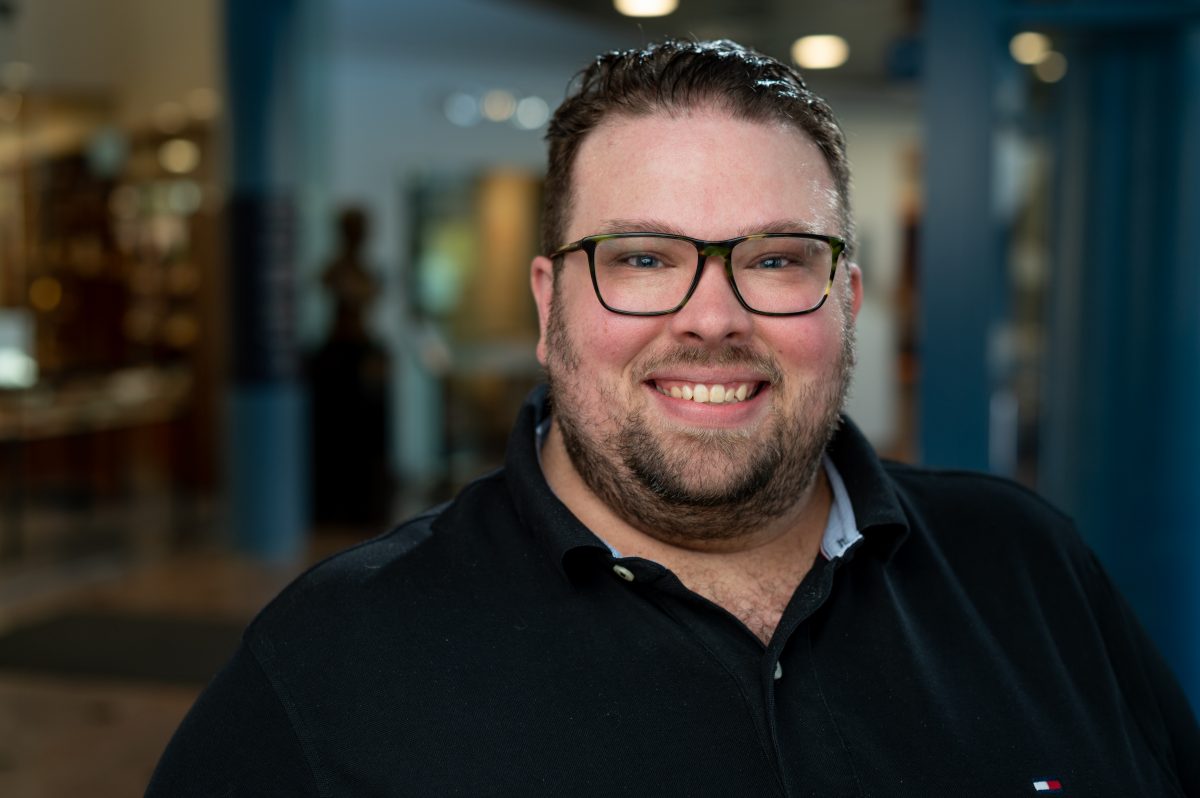 A man with brunette hair and facial hair wearing dark, tortoise shell glasses and a black polo smiles at the camera.